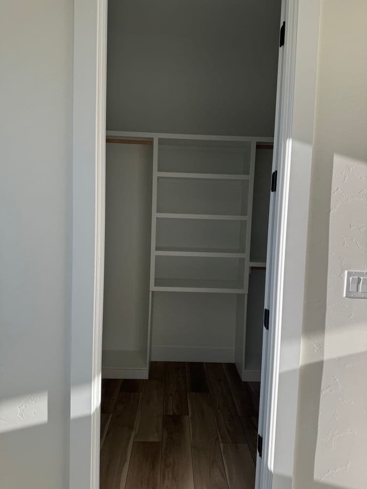 Empty closet with white shelving and wooden flooring, viewed from an open doorway.
