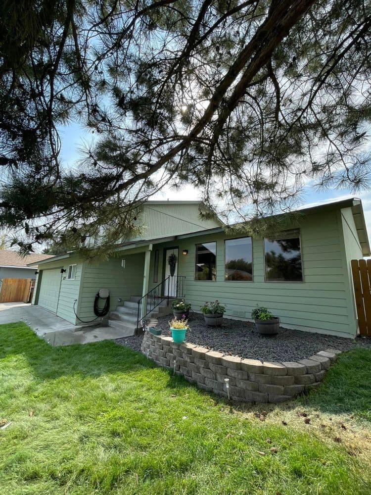 Green house with large windows, landscaped garden, and stone retaining wall under a blue sky.
