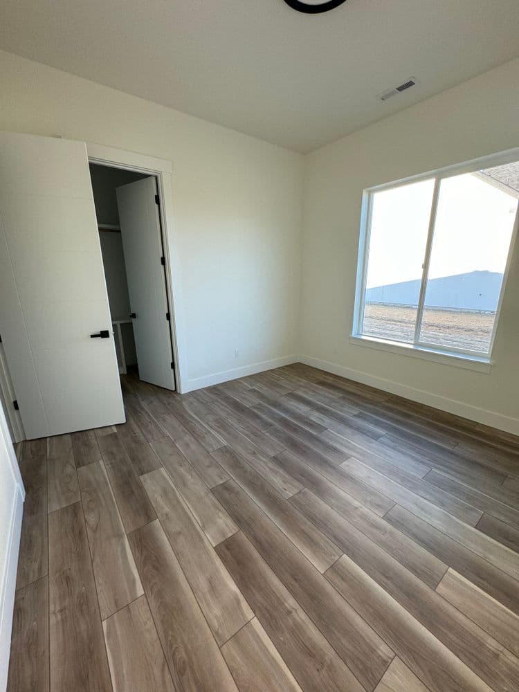 Empty bedroom with wooden flooring, bright natural light from large window, and open closet.